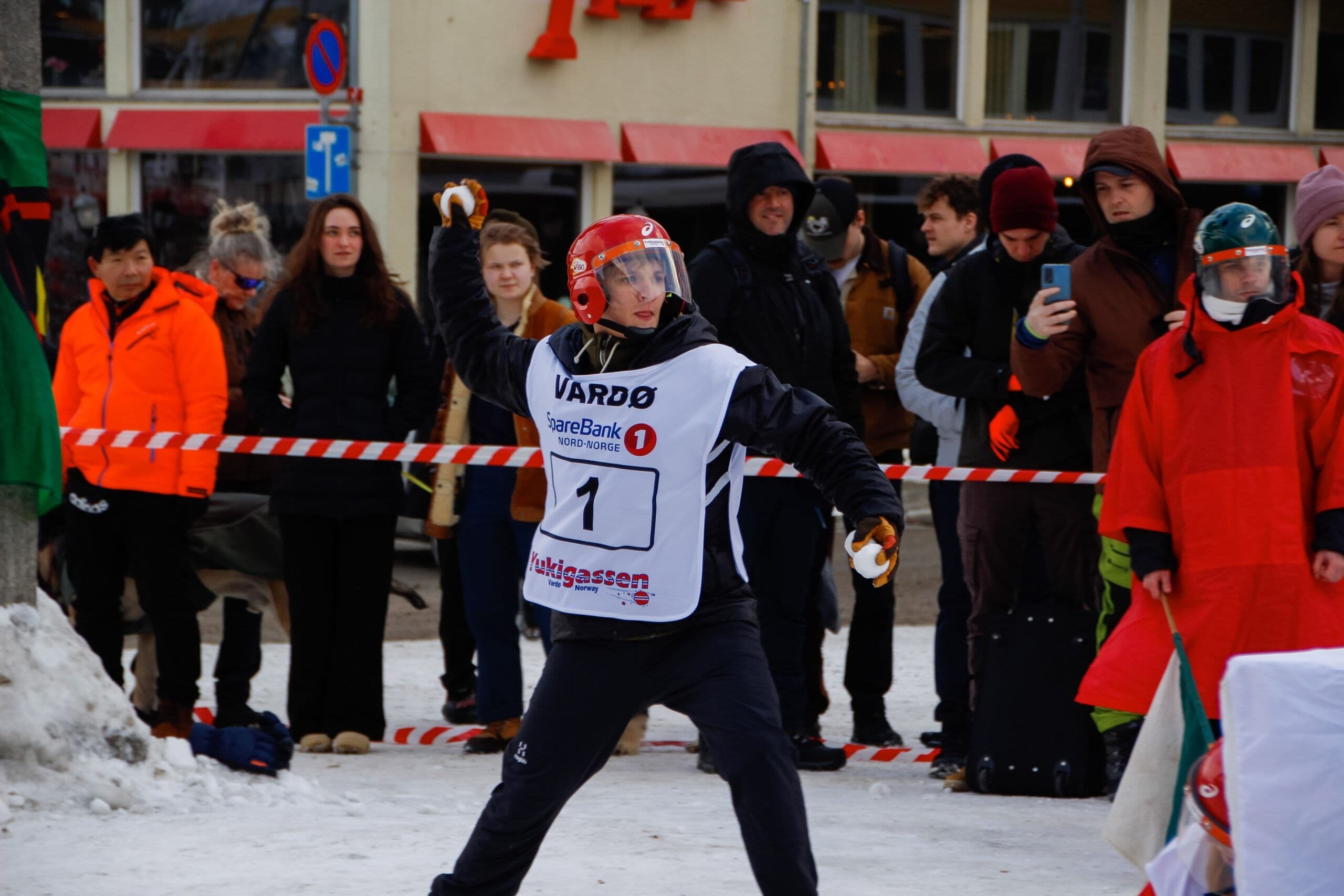 En utøver i verneutstyr og hjelm kaster en snøball under en utendørs vinterkonkurranse med publikum.