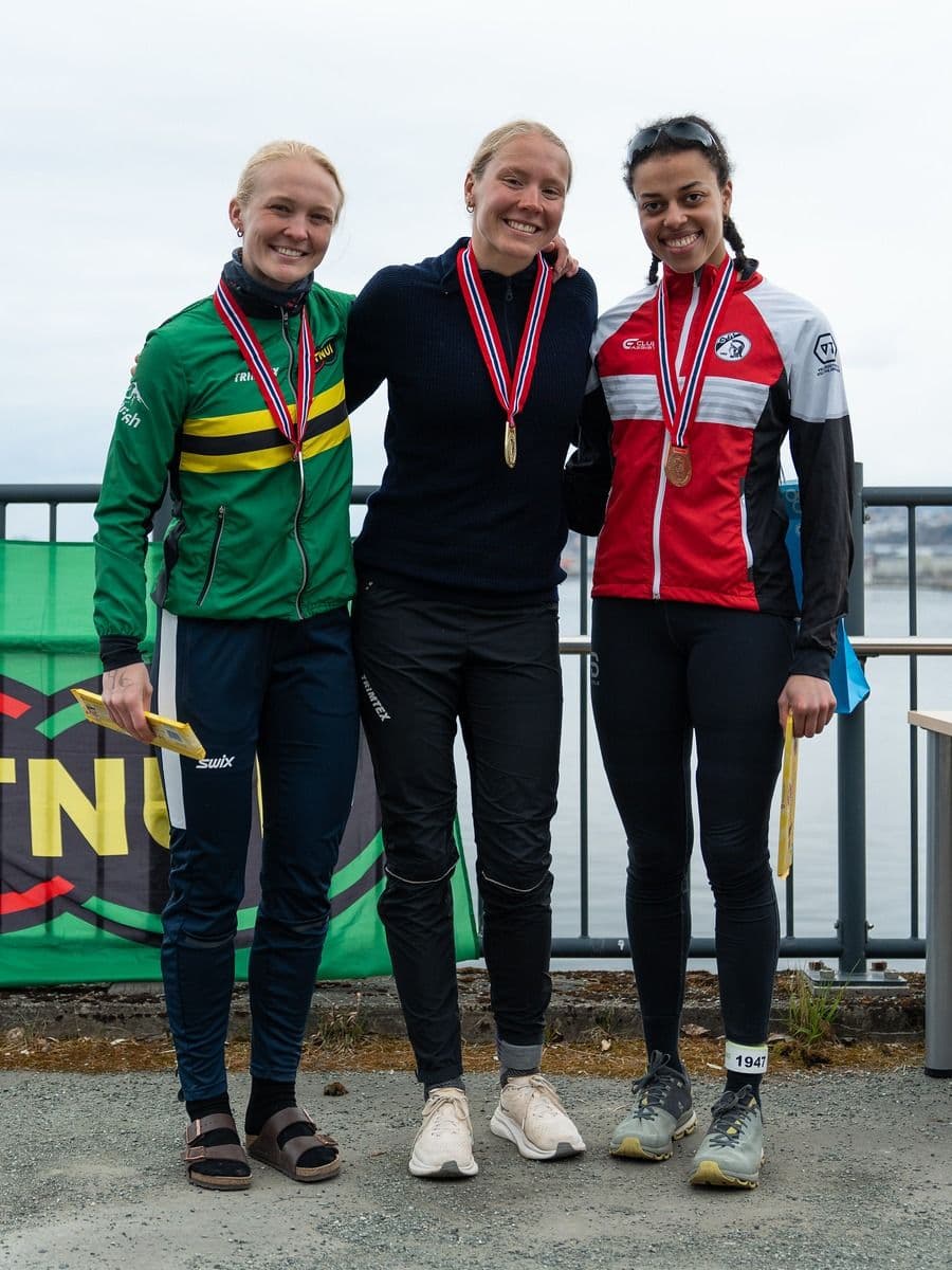 Tre kvinnelige utøvere med medaljer rundt halsen poserer på en premiepall ved vannet.