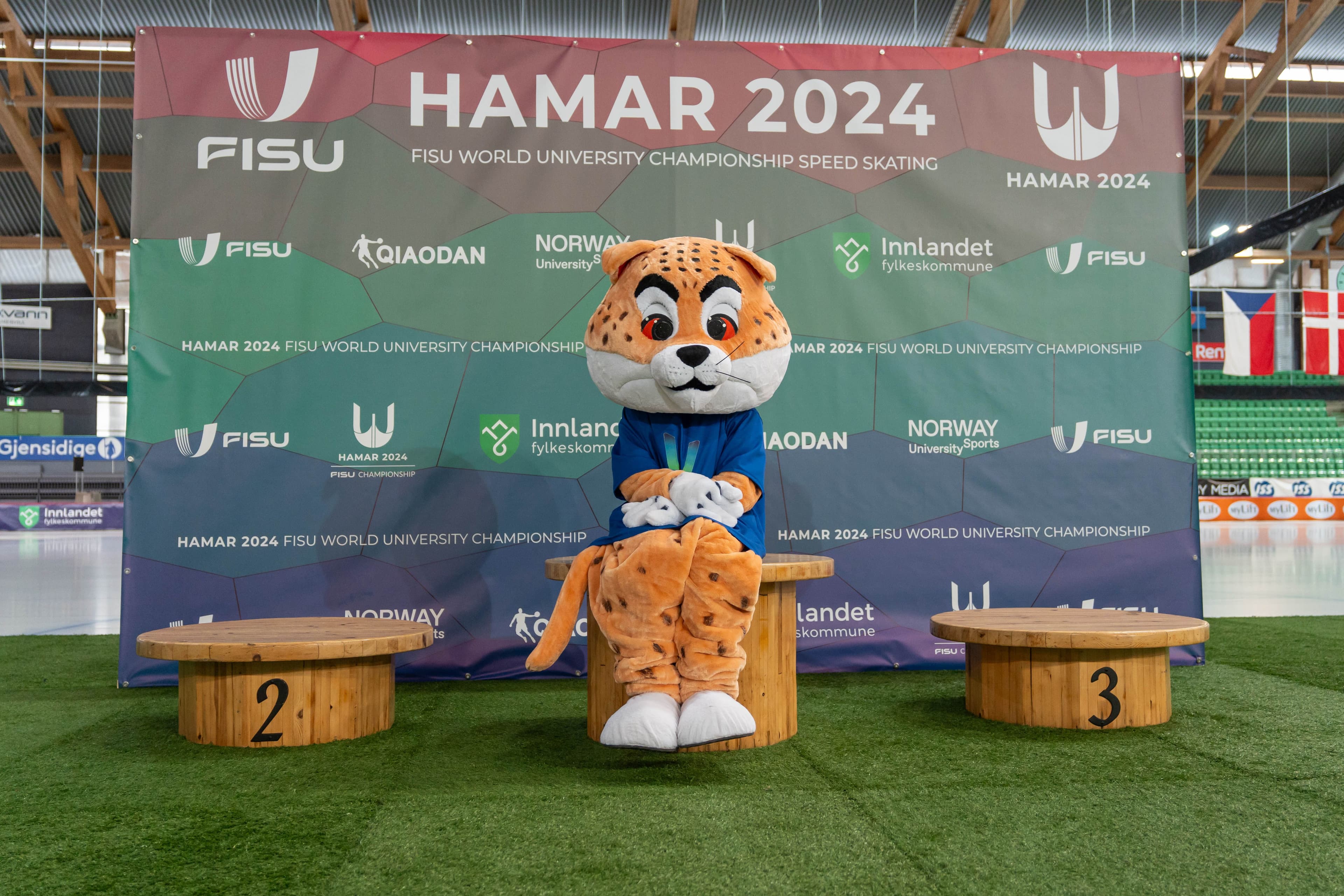 Maskoten, en gepard i blå trøye, sitter på seierspallen foran veggen for Hamar 2024 FISU World University Championship Speed Skating.