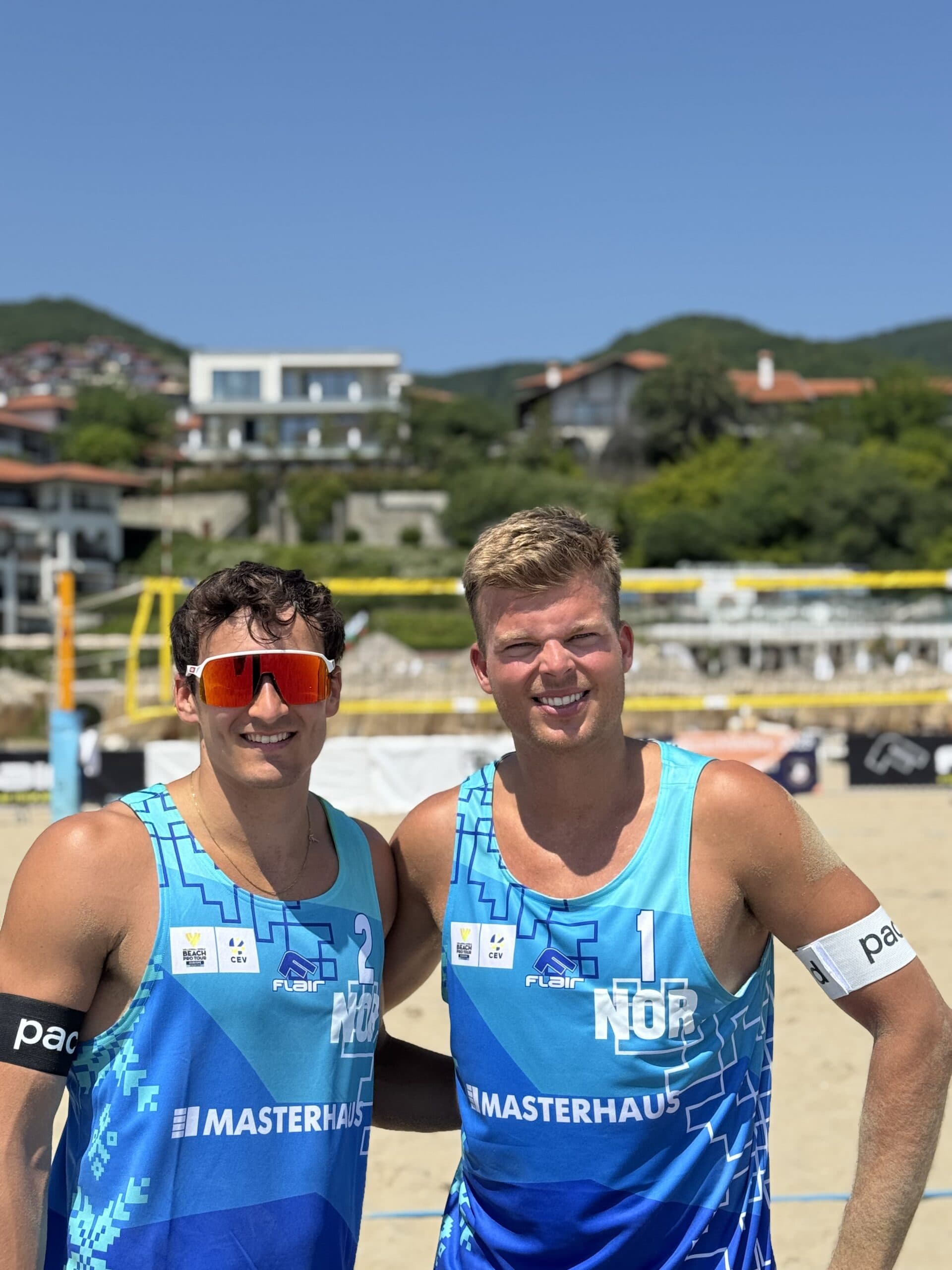 To mannlige sandvolleyballspillere i blå norske drakter poserer smilende på en strand.