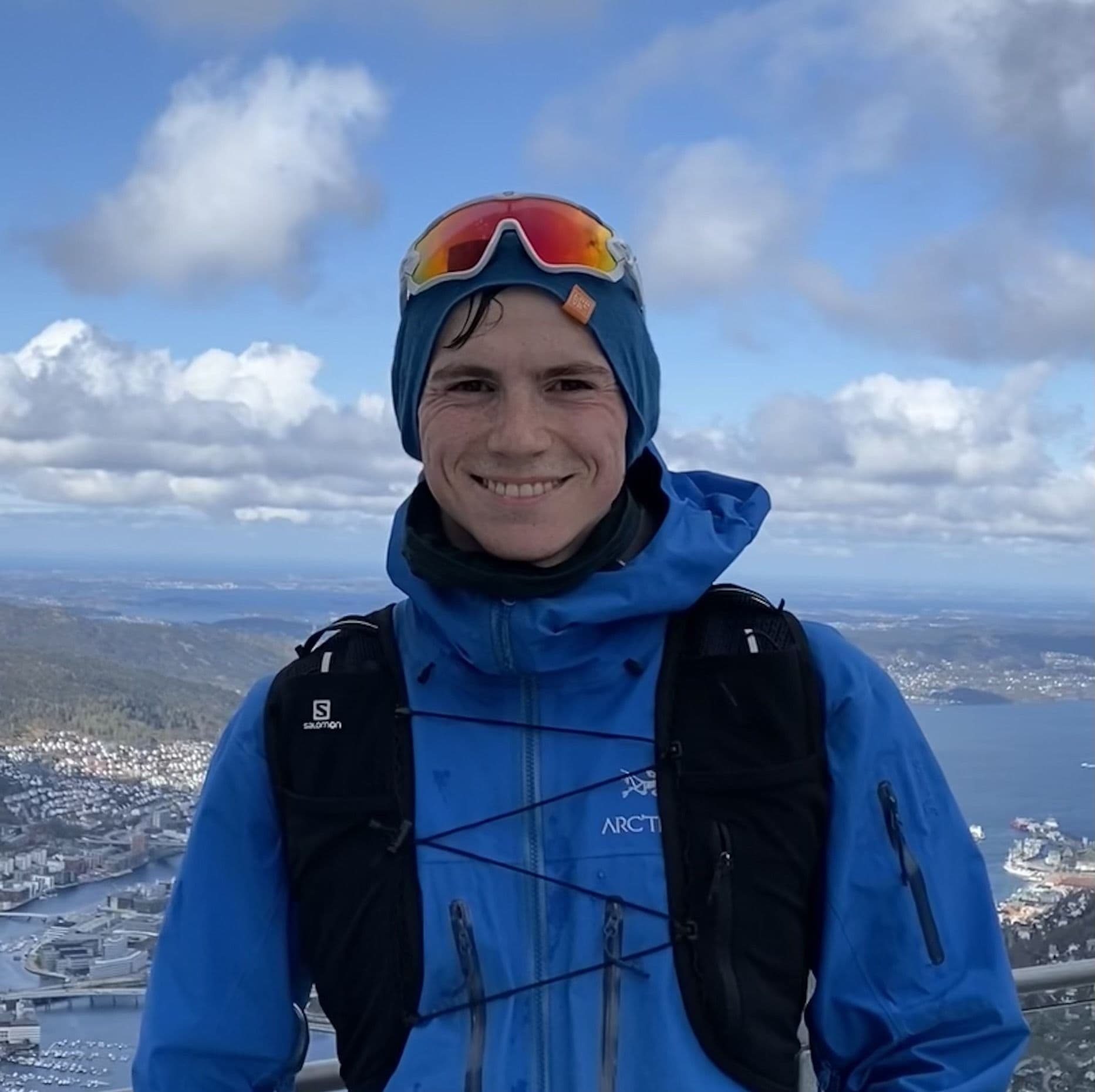 Portrett av en mann i blå jakke på en fjelltopp med utsikt over en by og fjord.