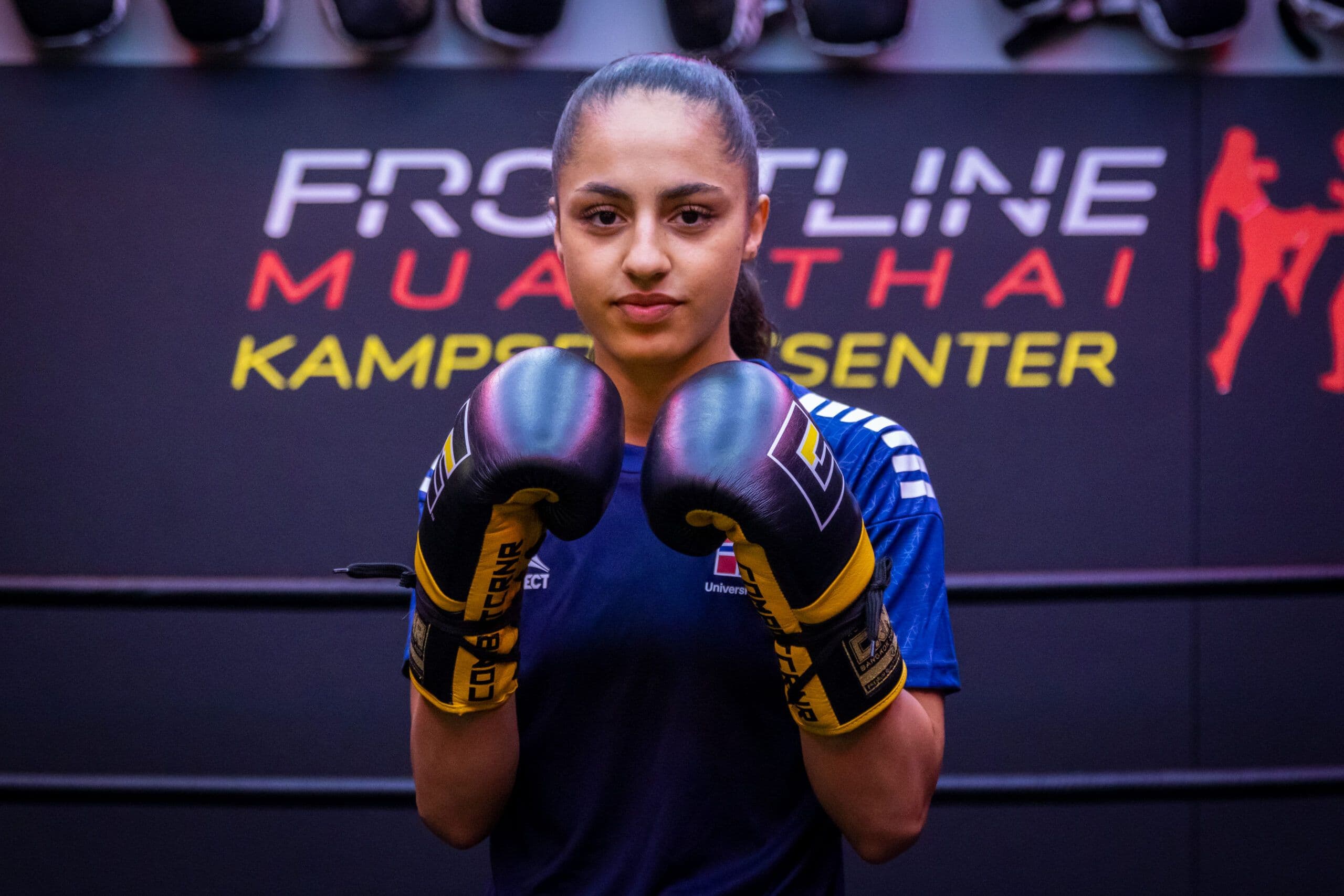 En ung utøver med boksehansker poserer foran en vegg med teksten «Frontline Muay Thai».