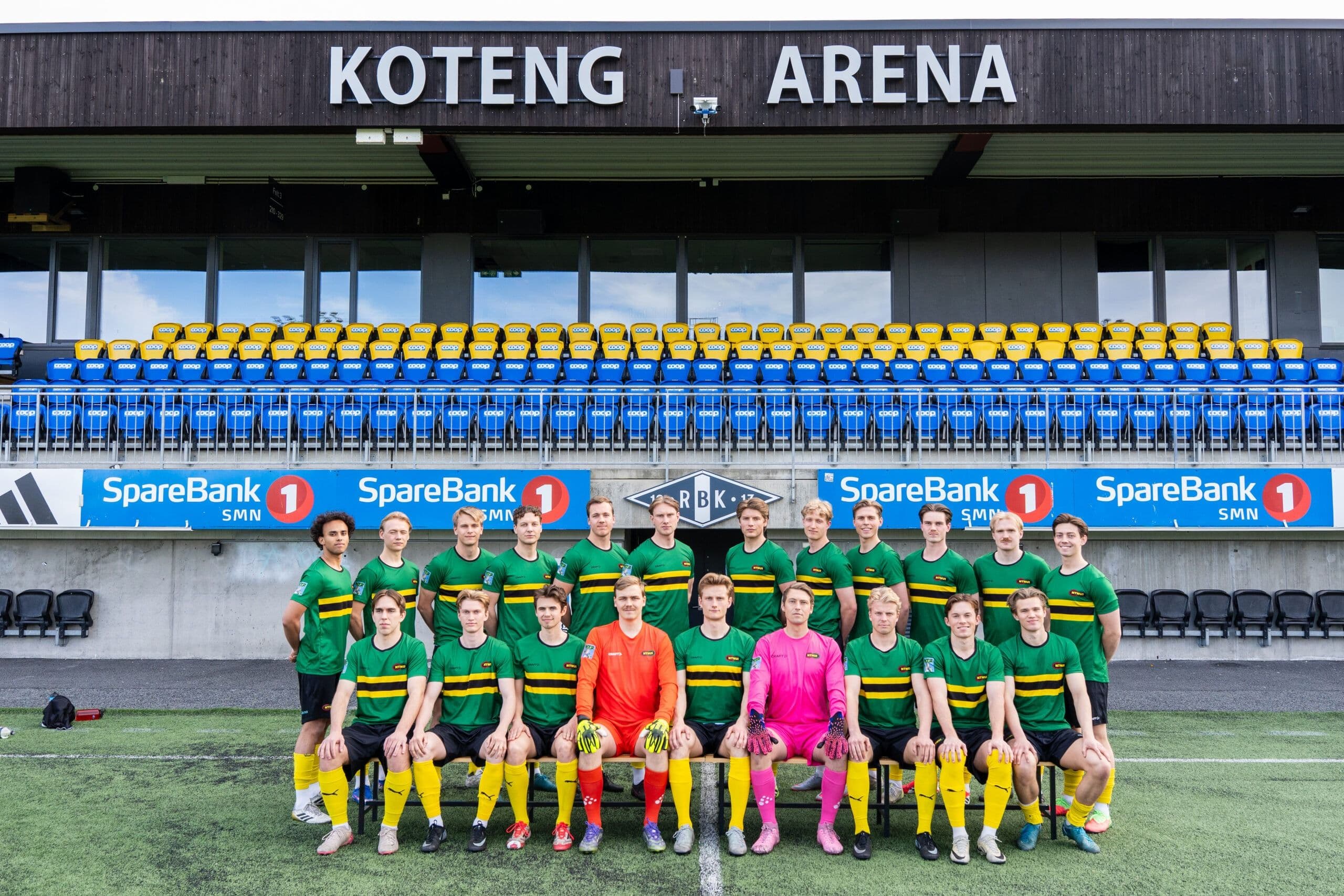 Lagbilde av et fotballag i grønne og gule drakter foran tribunen på Koteng Arena.