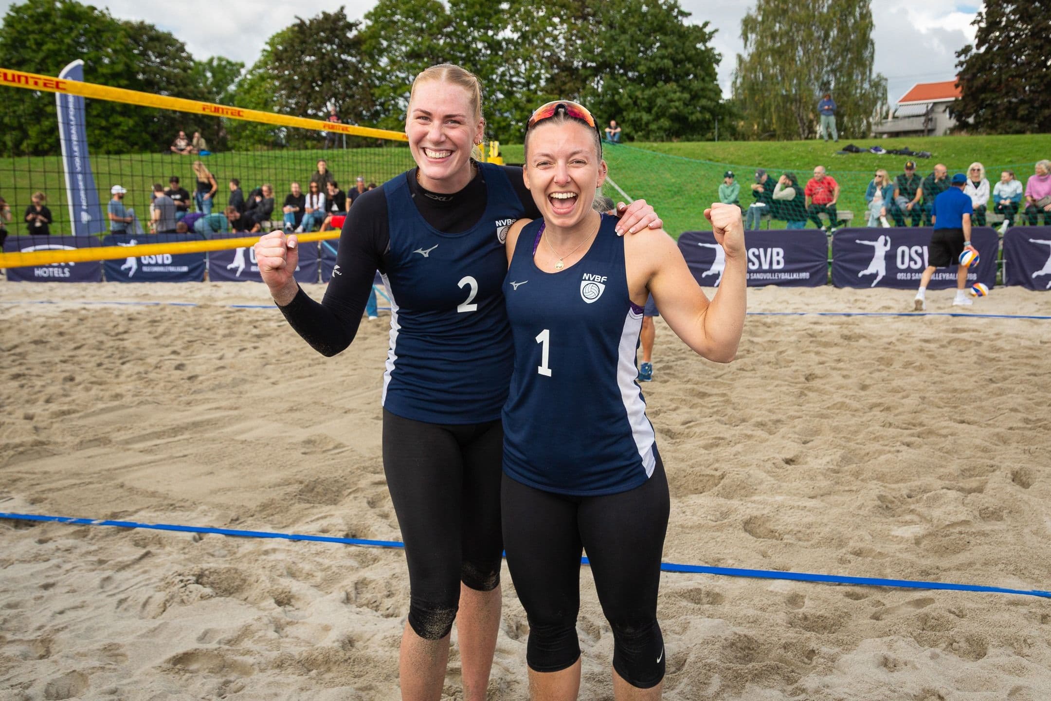 To kvinnelige sandvolleyballspillere jubler sammen ved nettet på en sandbane.
