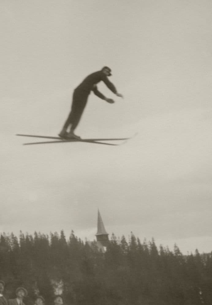 Svart-hvitt-foto av en skihopper høyt i lufta over skog med kirketårn i bakgrunnen.