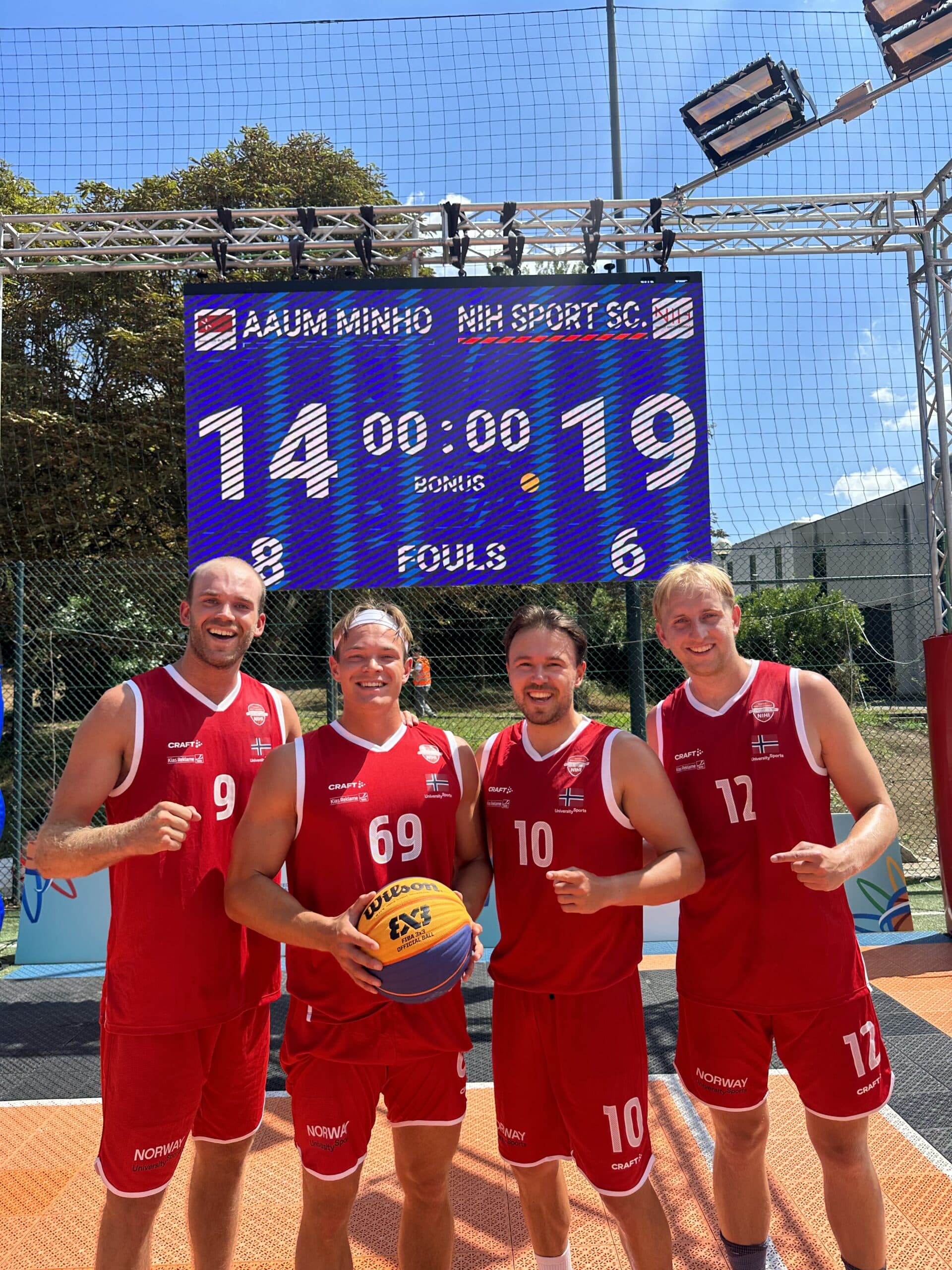 Fire mannlige basketballspillere i røde Norway-drakter poserer foran en resultattavle på en utebane.