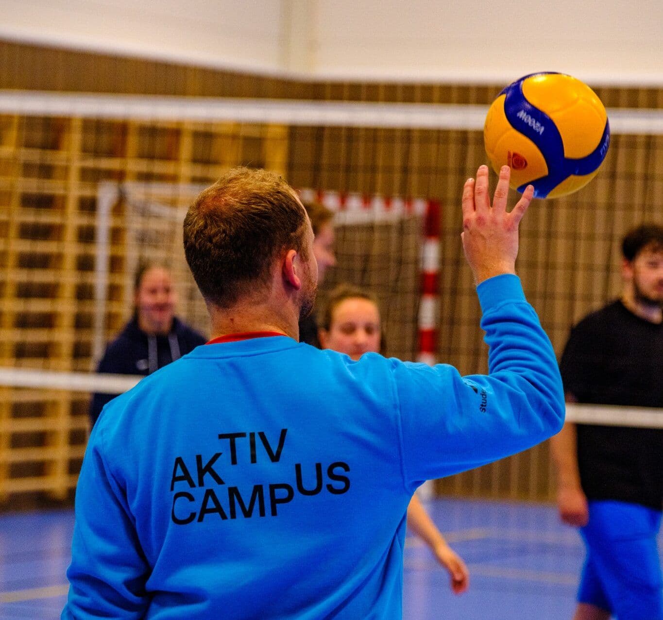 En mann i blå "Aktiv Campus"-genser slår volleyballserve mot nettet i en gymsal.