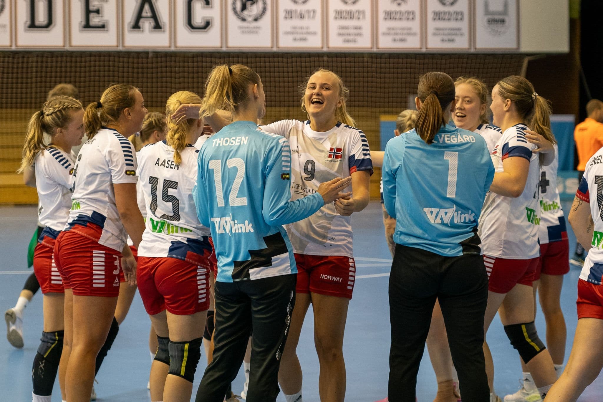 Et kvinnelig håndballag i norske landslagsdrakter smiler og feirer sammen i en idrettshall.