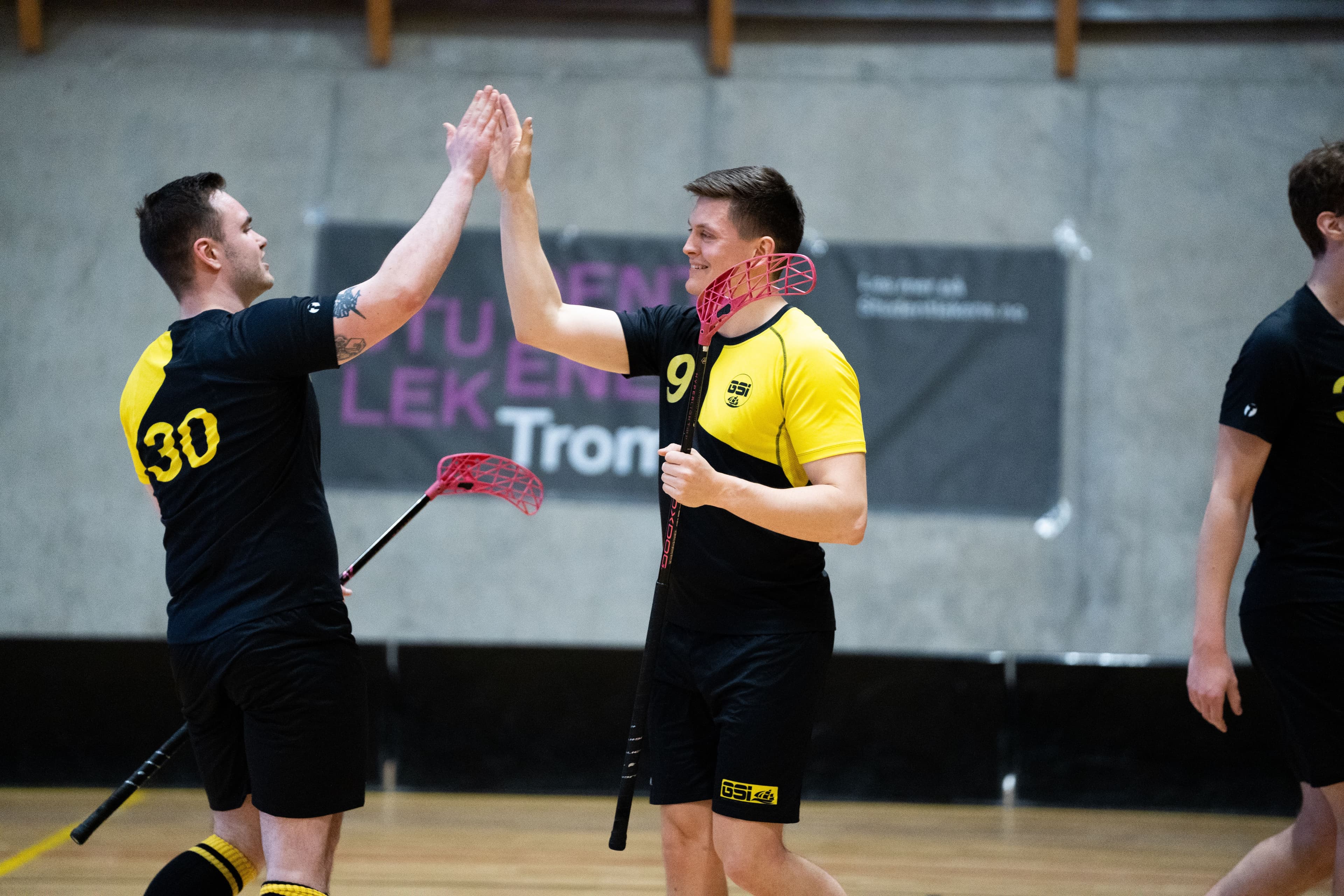 To mannlige innebandyspillere i gult og svart gir hverandre high five i en idrettshall.