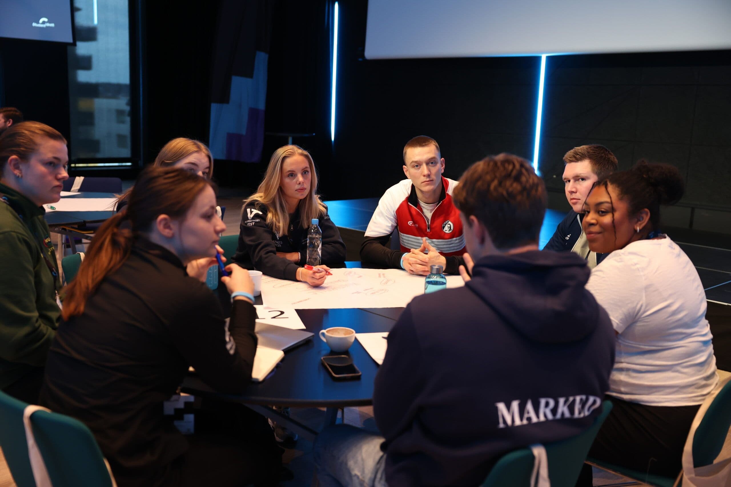En gruppe studenter sitter rundt et bord og diskuterer under en workshop.