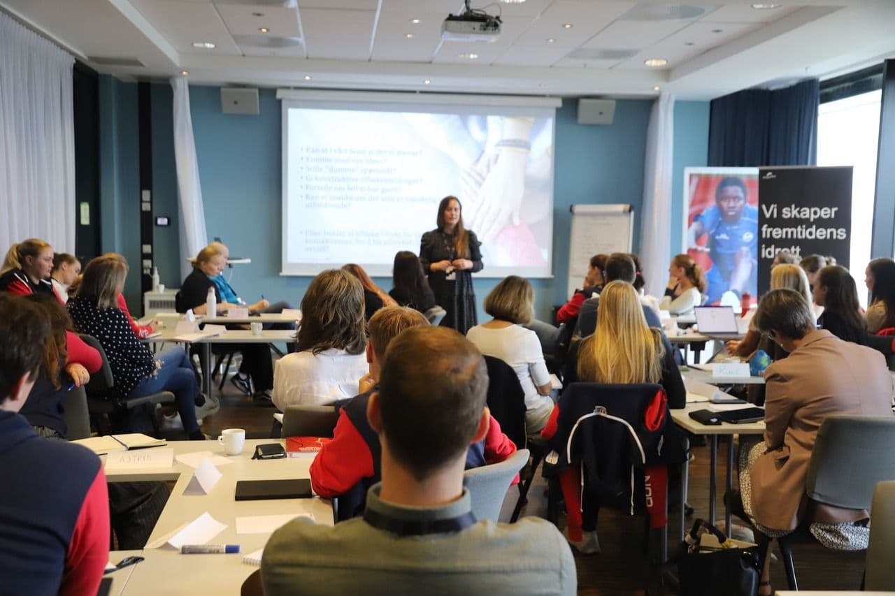 En person holder et foredrag foran en gruppe deltakere i et konferanserom, med rollup med teksten Vi skaper fremtidens idrett.