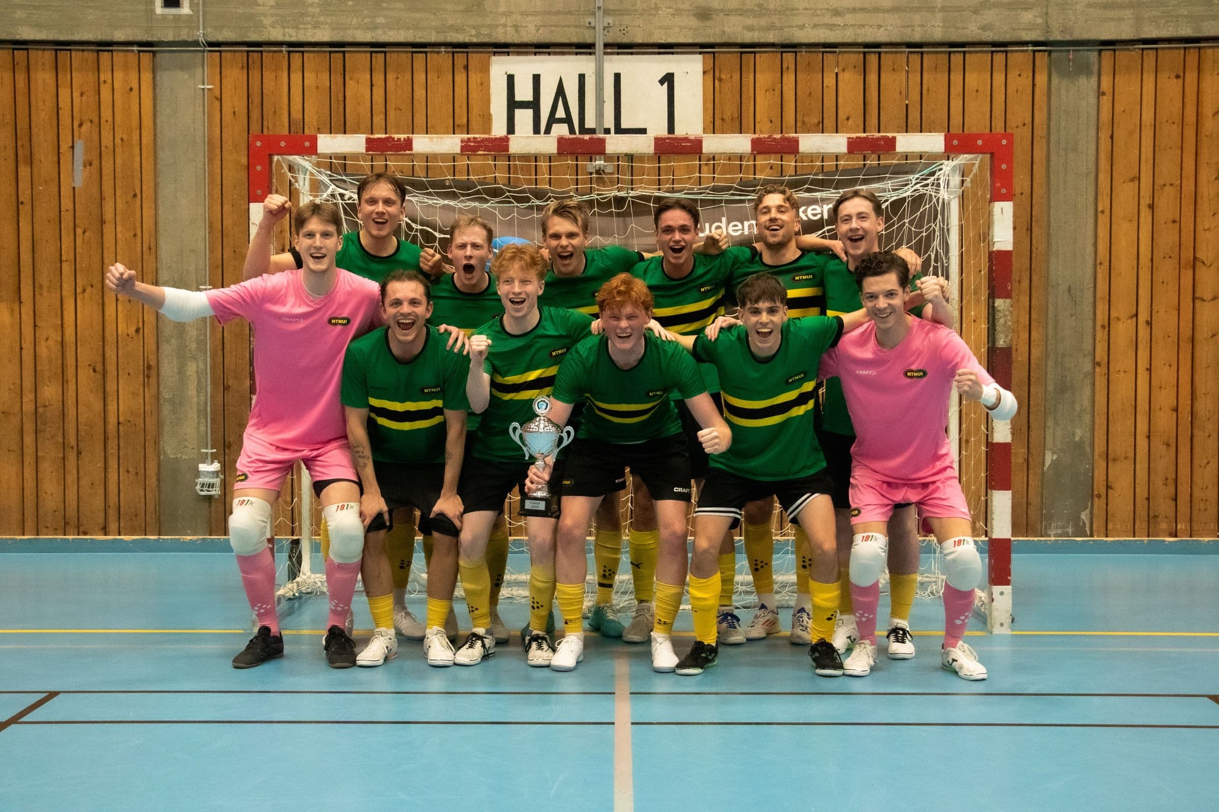 Et futsallag i grønne og rosa drakter jubler med en pokal foran målet i en idrettshall.