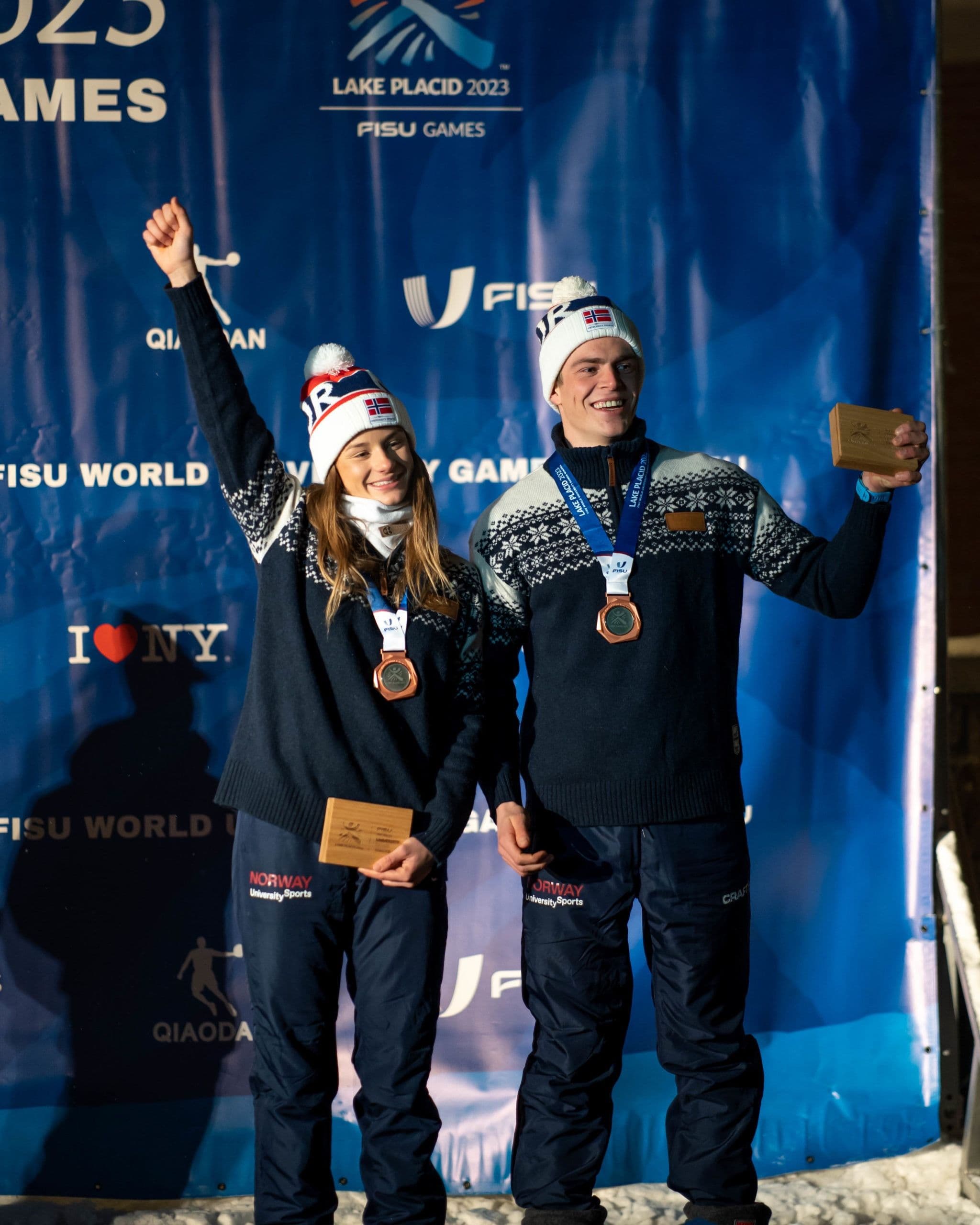 To utøvere med medaljer jubler med armene i været under FISU-lekene i Lake Placid 2023.