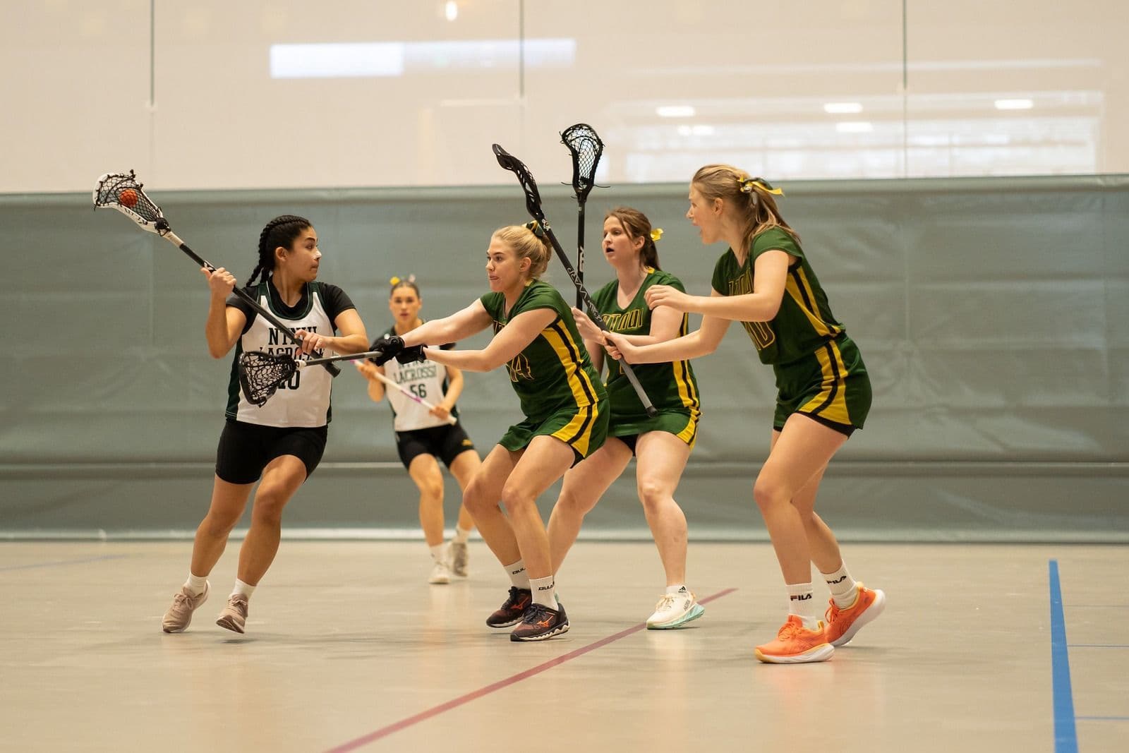 Spillere i lacrosse i grønne og hvite drakter kjemper om ballen med køller i en idrettshall.