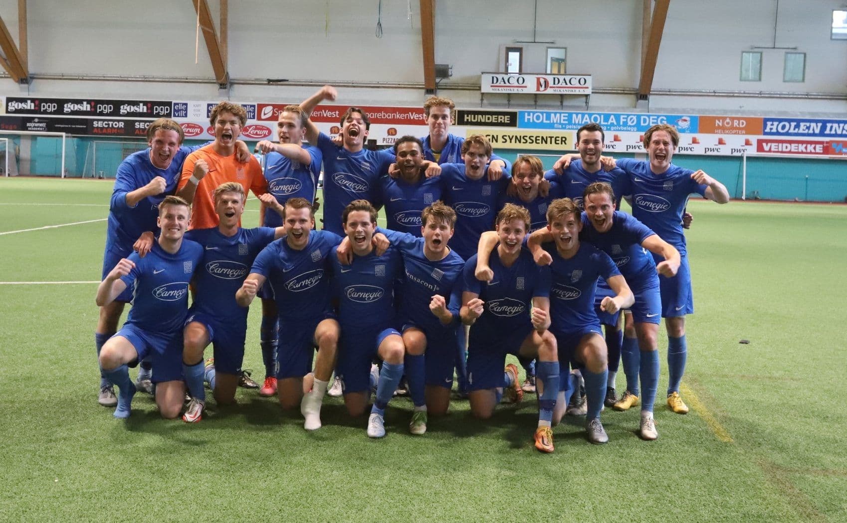 Et jublende futsallag i blå drakter poserer sammen på kunstgress i en idrettshall.