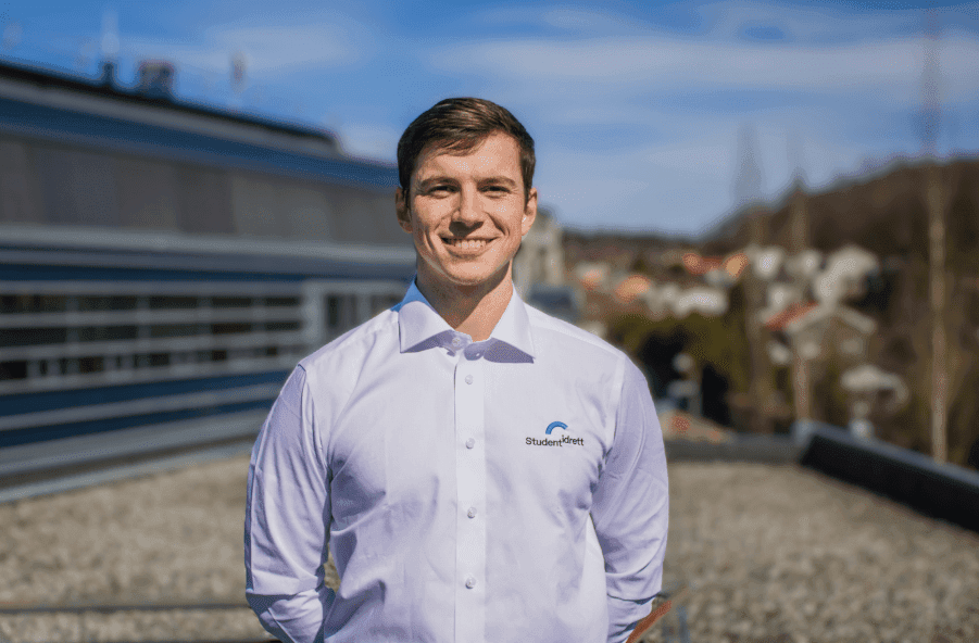Portrett av en smilende mann i hvit skjorte utendørs på et tak med utsikt over landskap.
