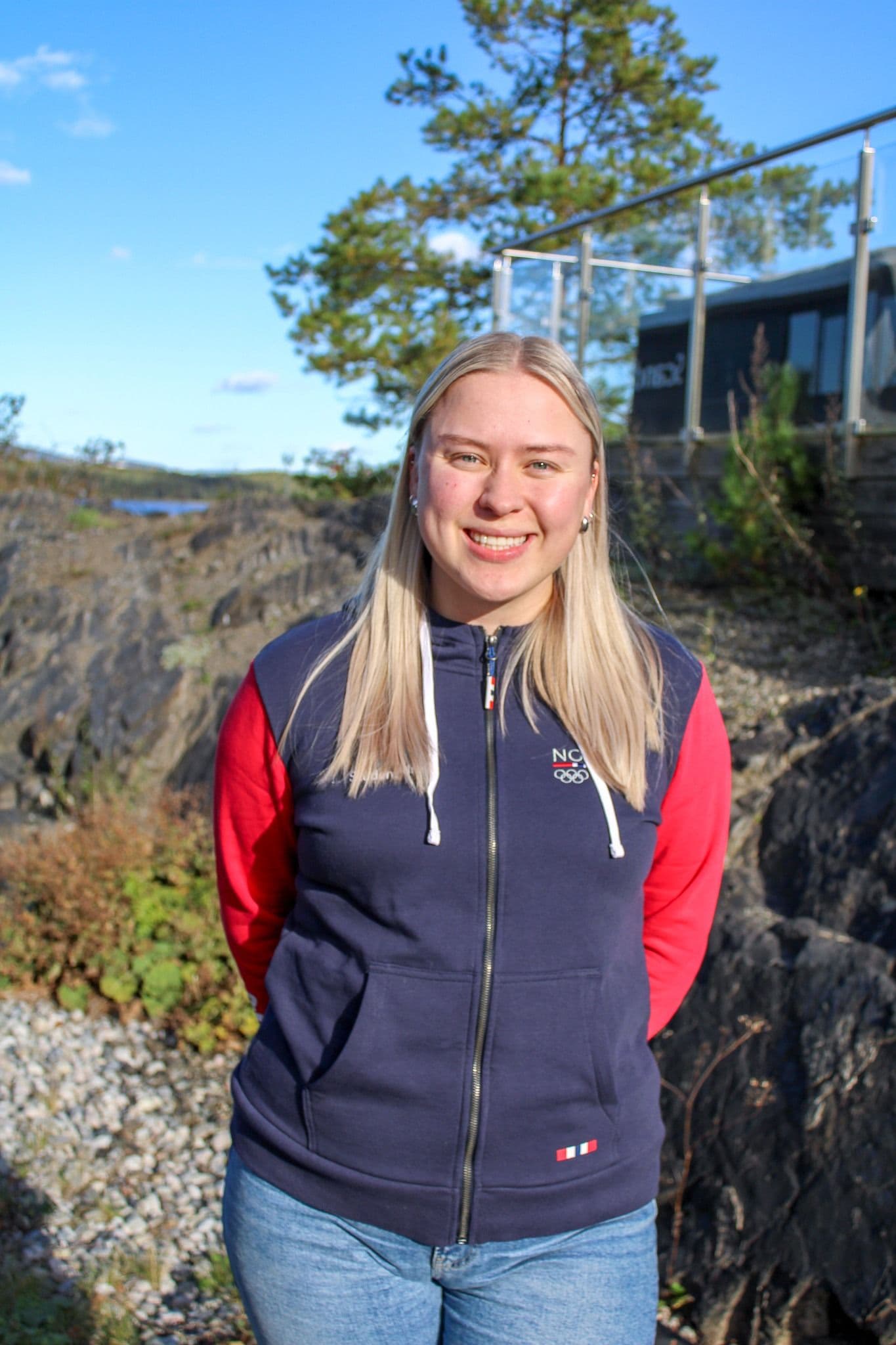 Portrett av en smilende ung kvinne i marineblå vest med Norge-logo, ute i naturen.