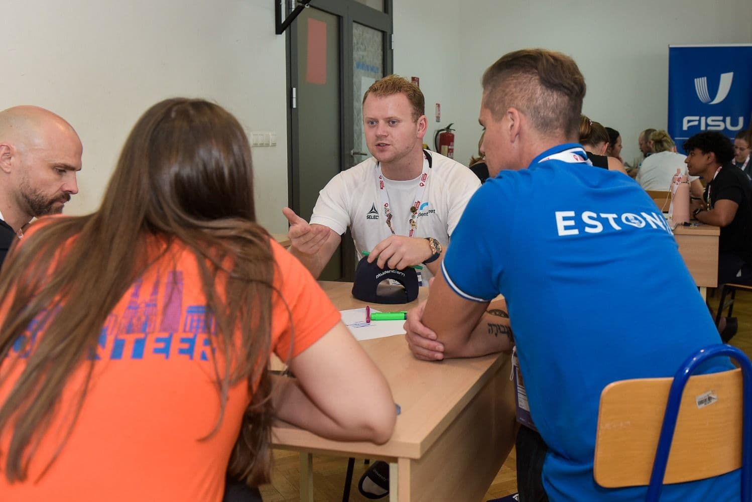 Flere personer sitter i samtale rundt et bord ved et FISU-arrangement, én i trøye merket Estonia.