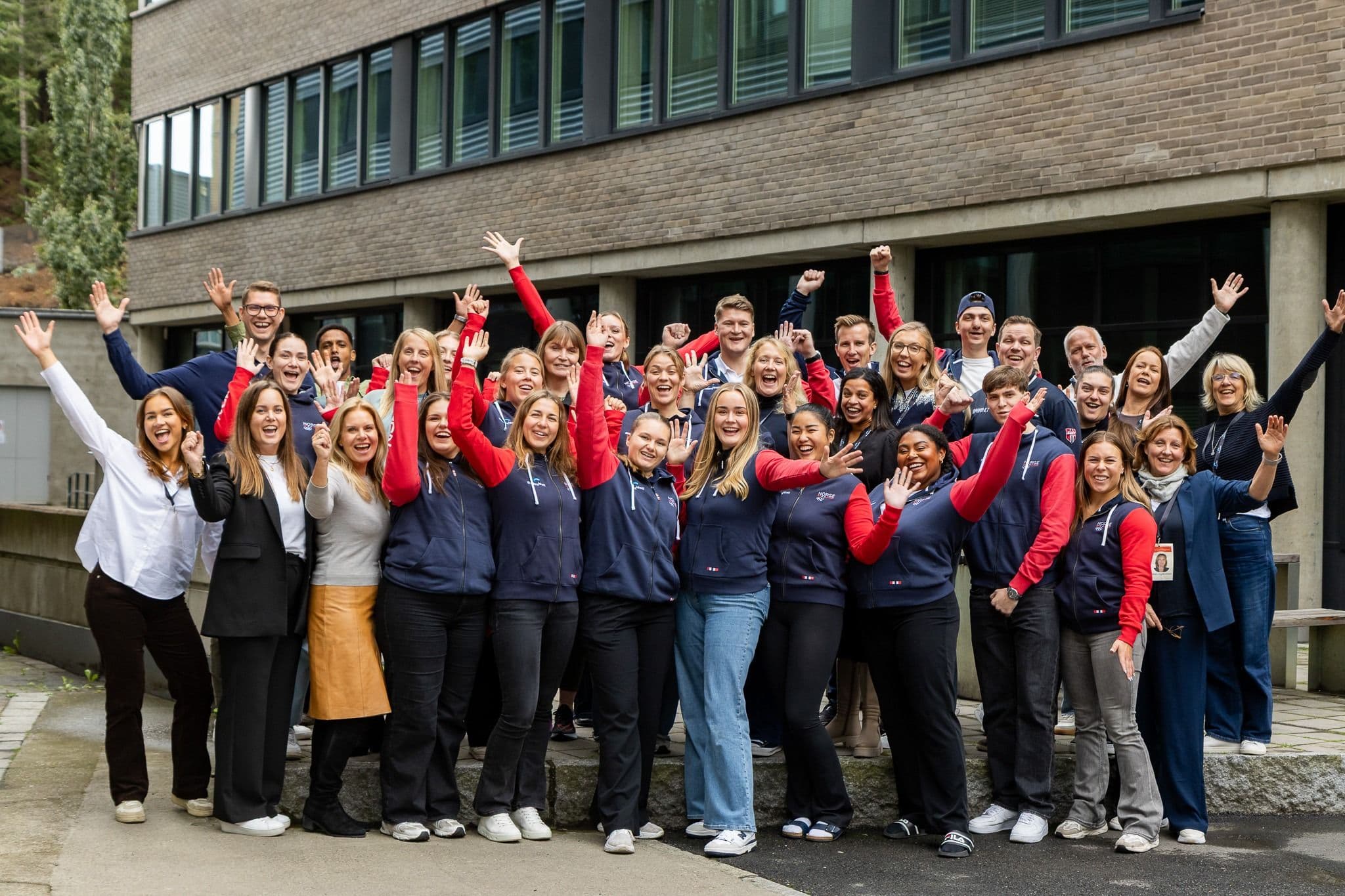 En stor gruppe studenter i Norge-antrekk jubler med armene i været utenfor et bygg.