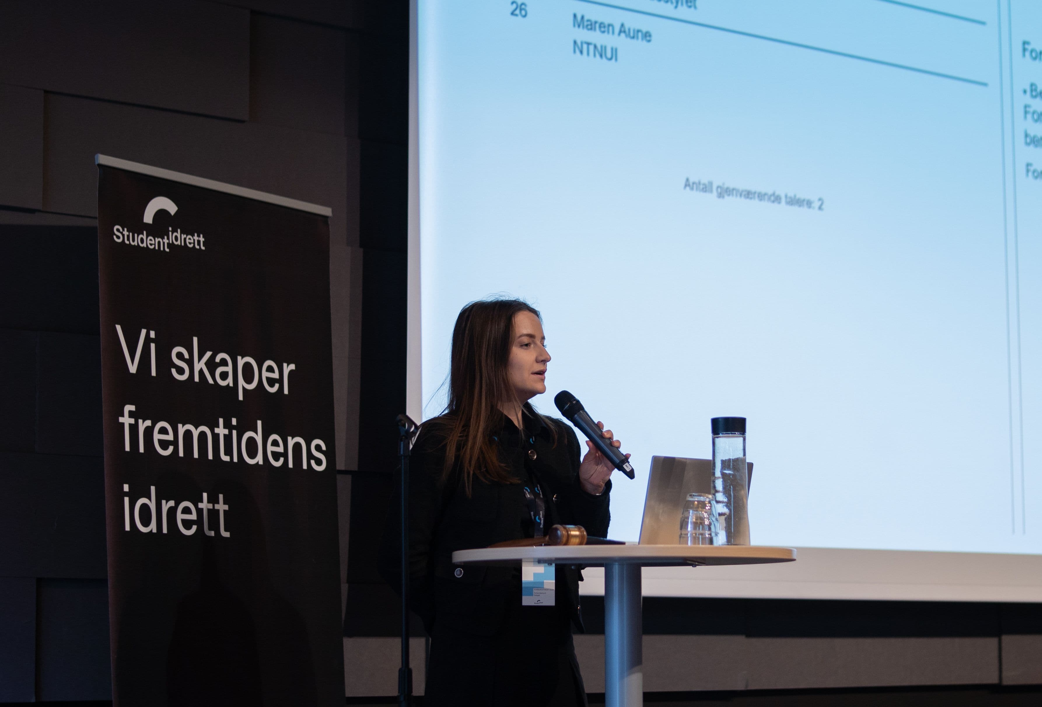 En kvinne holder innlegg med mikrofon på en scene ved siden av en Studentidrett-beachflag med teksten Vi skaper fremtidens idrett.