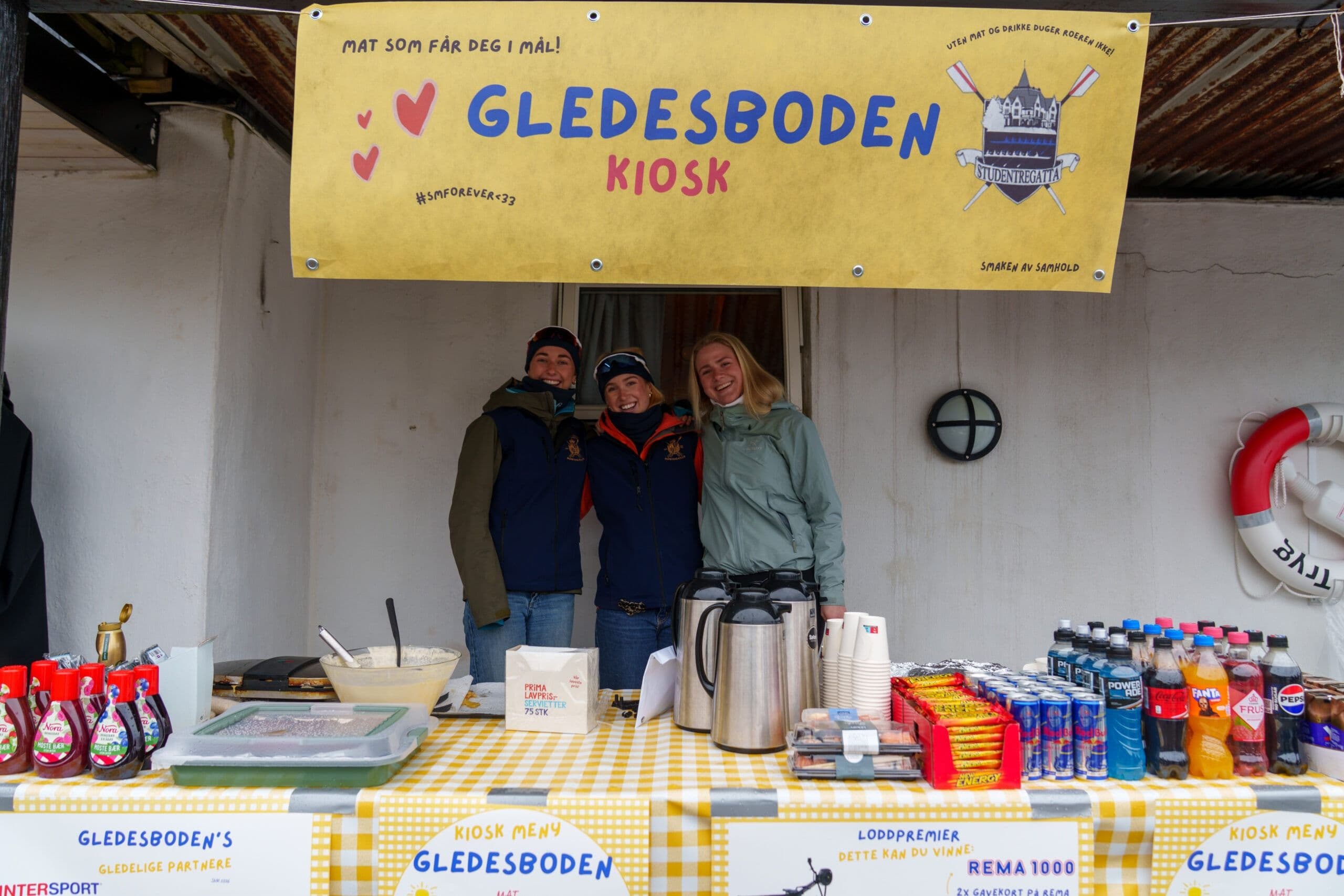 Tre unge personer står bak disken i «Gledesboden Kiosk» med kaffe og varer.