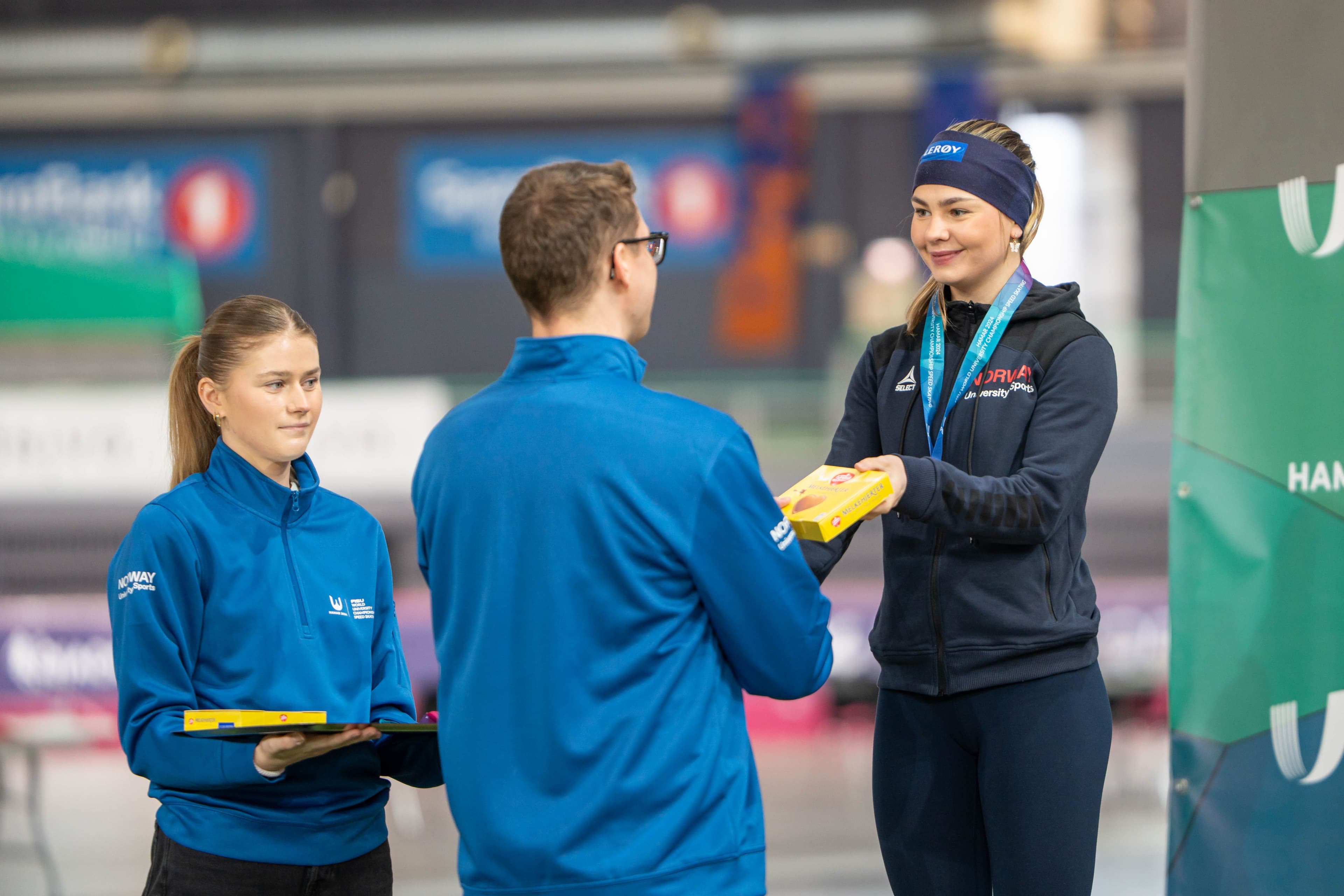 En utøver med medalje mottar en gave fra to arrangører i blå Studentidrett-jakker.