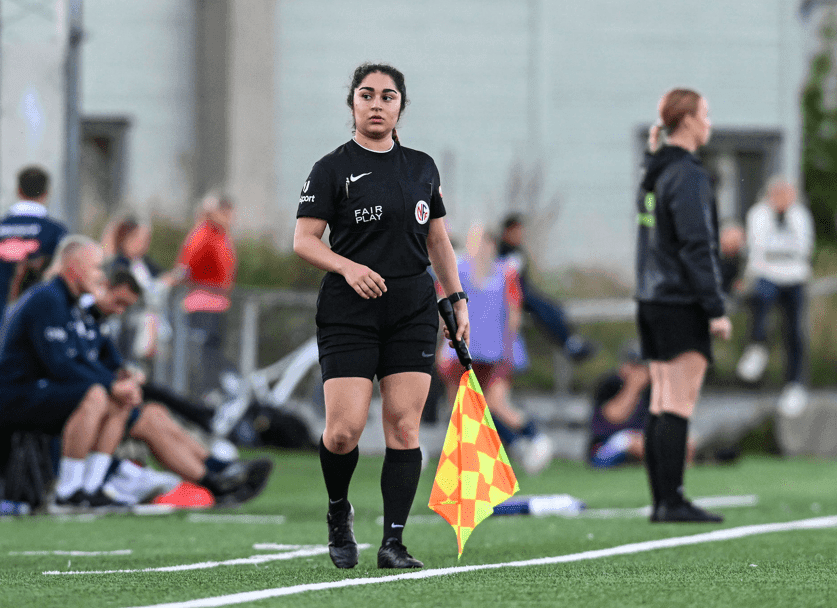 En kvinnelig assistentdommer med linjemannsflagg langs sidelinjen på en fotballbane.