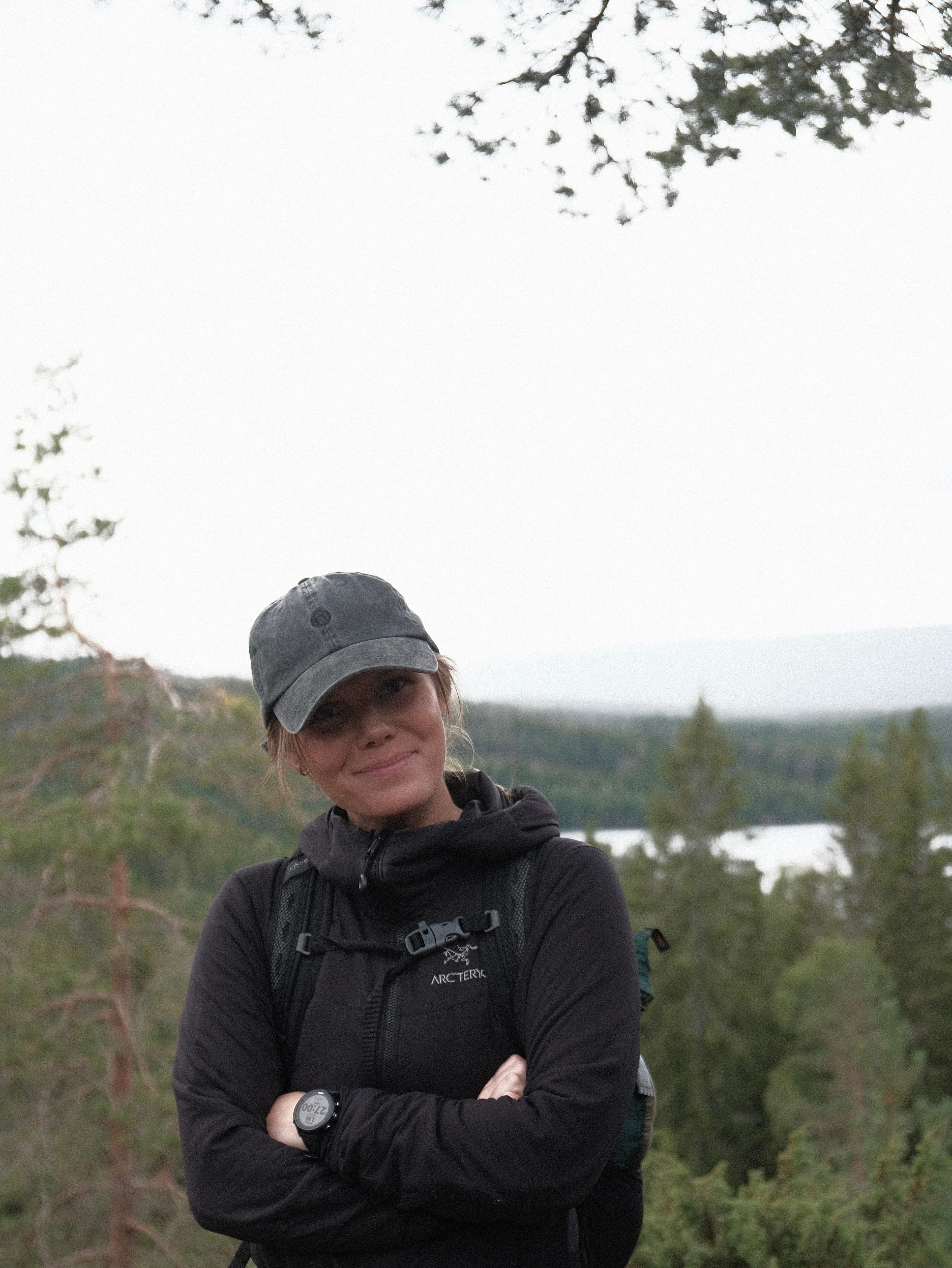 Portrett av en kvinne med caps og turjakke foran skog og et vann.