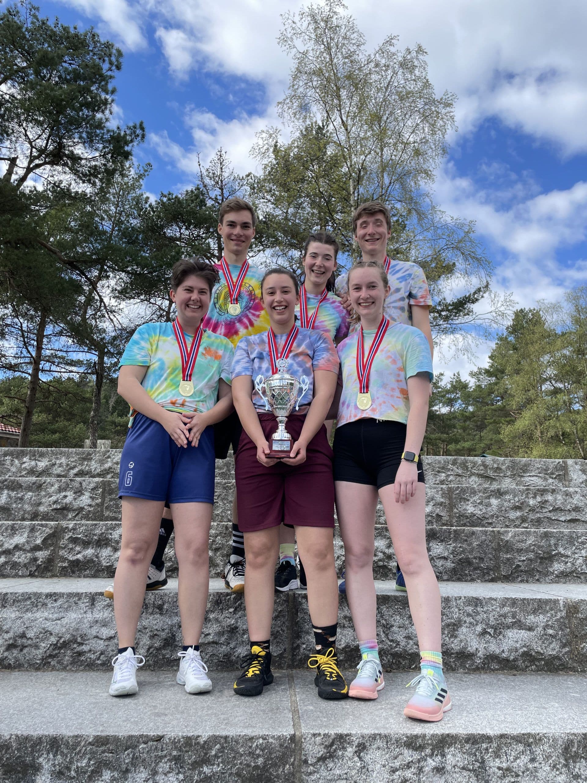 Seks studenter med medaljer poserer med en pokal på en steintrapp i solskinn.