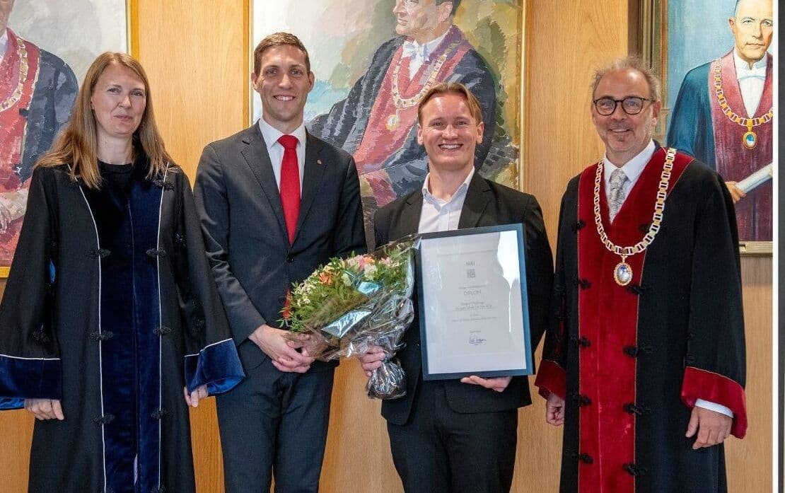 Fire personer poserer under en prisutdeling, en holder blomster og diplom mens en rektor i kappe og kjede står ved siden.