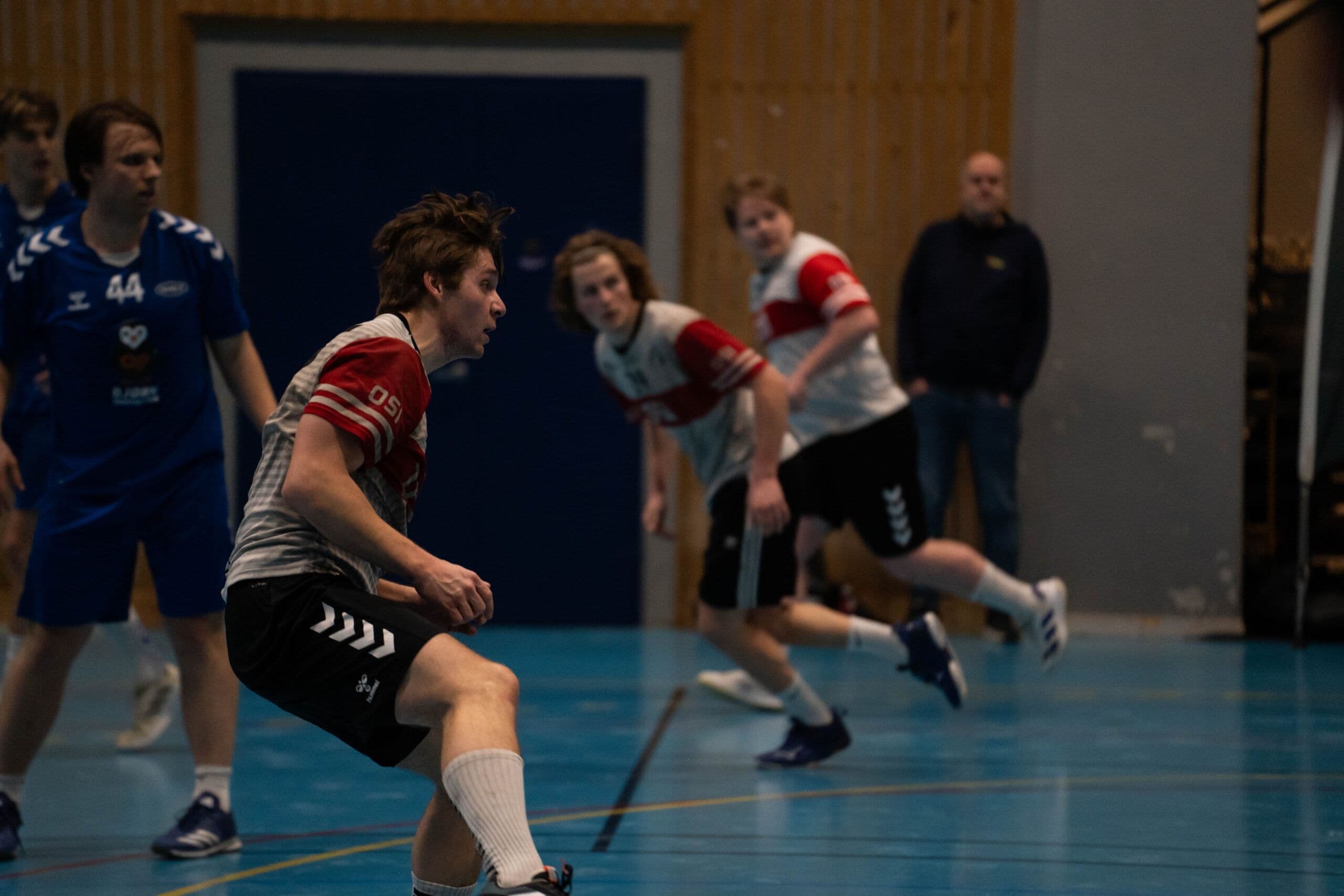 En håndballspiller i rød og grå drakt fører ballen under en kamp i en idrettshall.