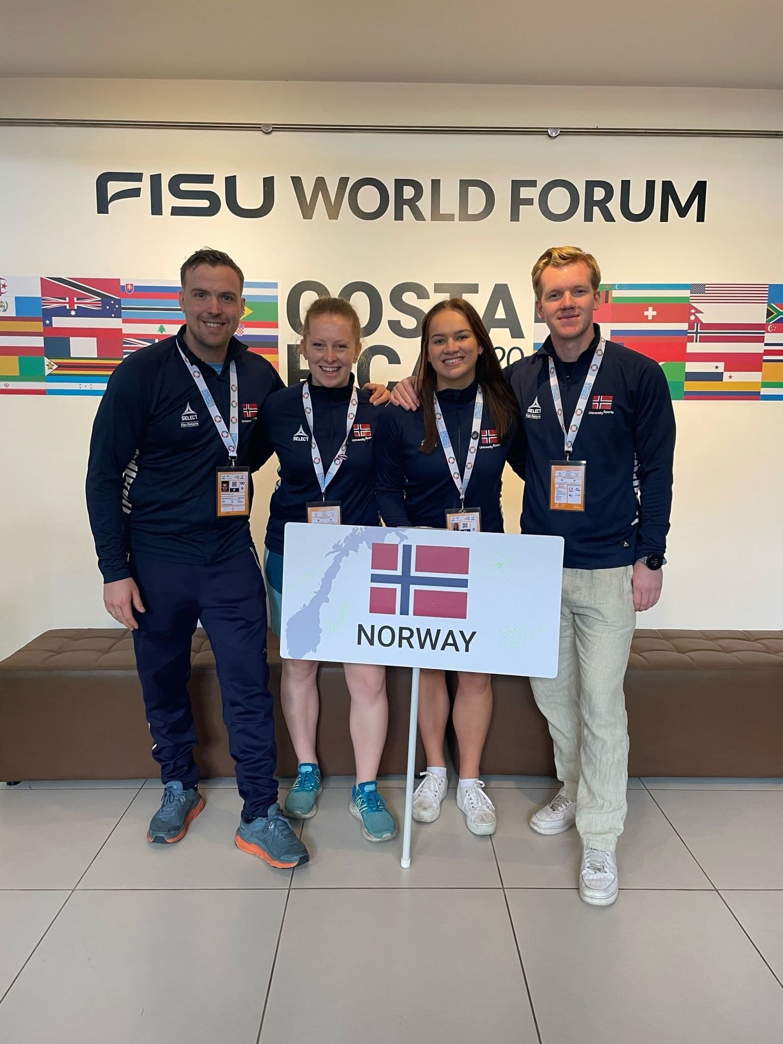 Fire personer i norske jakker poserer med et Norway-skilt foran en vegg med teksten FISU World Forum.