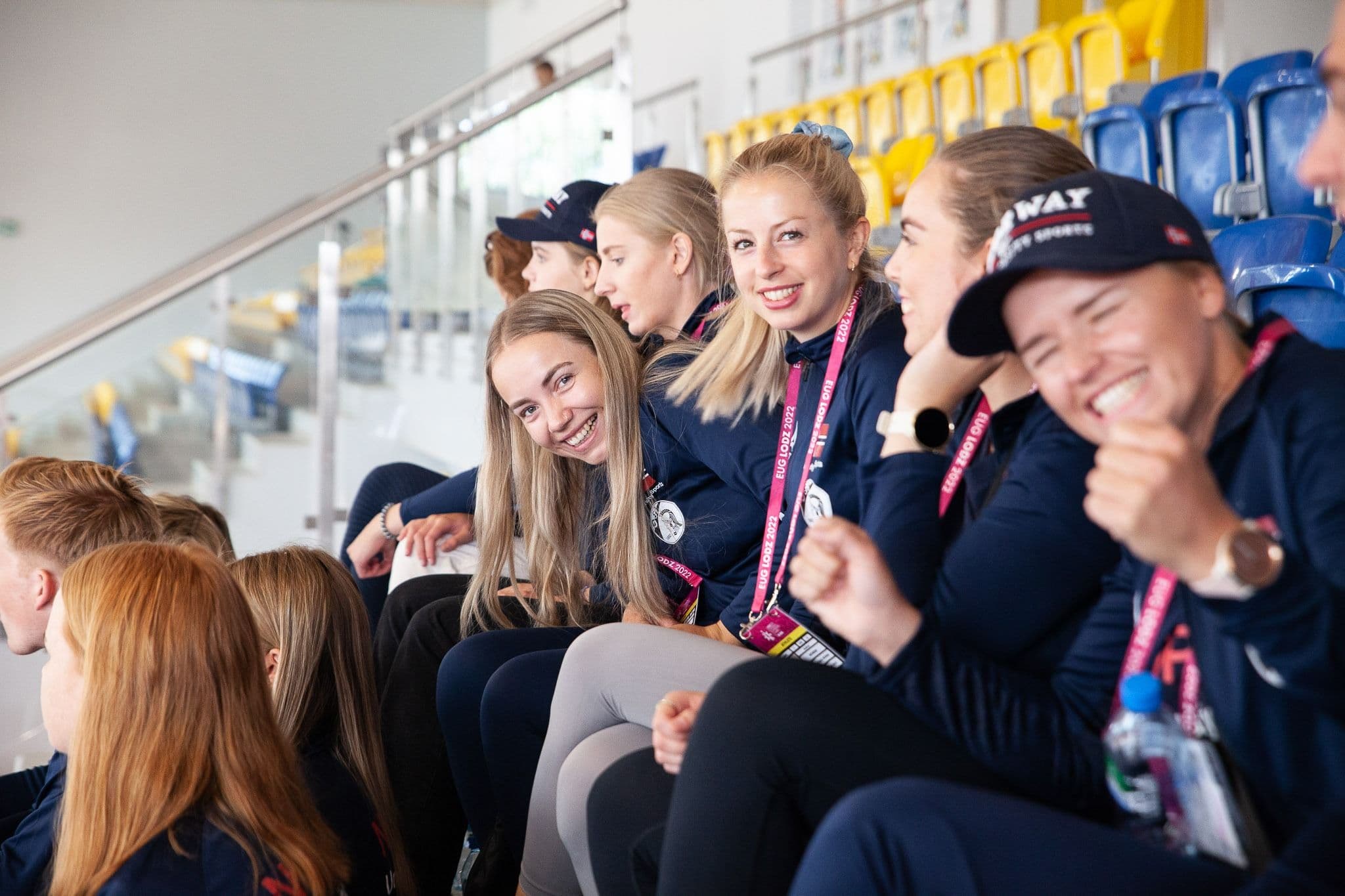 Smilende utøvere i norske landslagsjakker sitter på tribunen og følger en konkurranse.