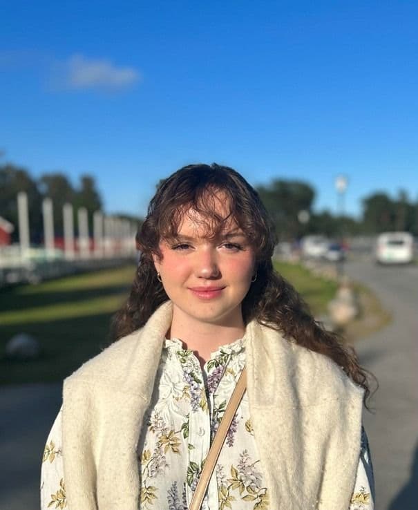 Portrett av en ung kvinne i blomstrete bluse og hvit vest utendørs med blå himmel i bakgrunnen.
