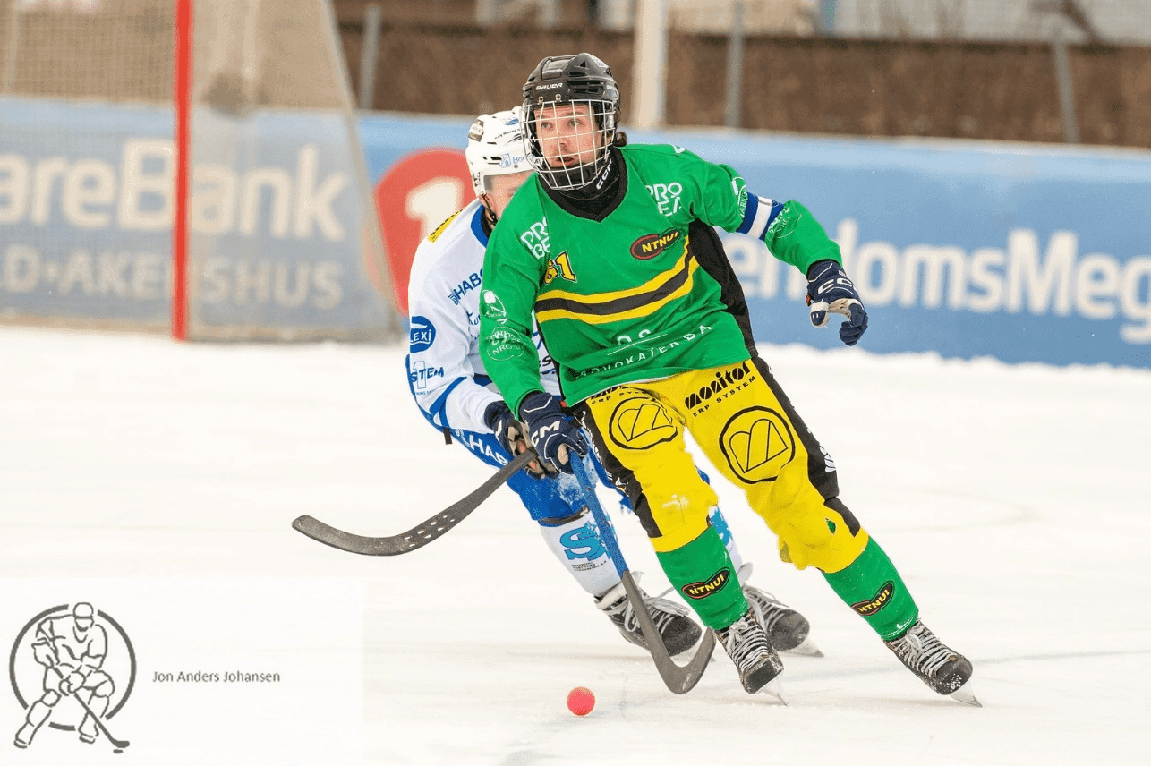 En bandyspiller i grønn og gul drakt fører ballen på isen forbi en motspiller under en kamp.